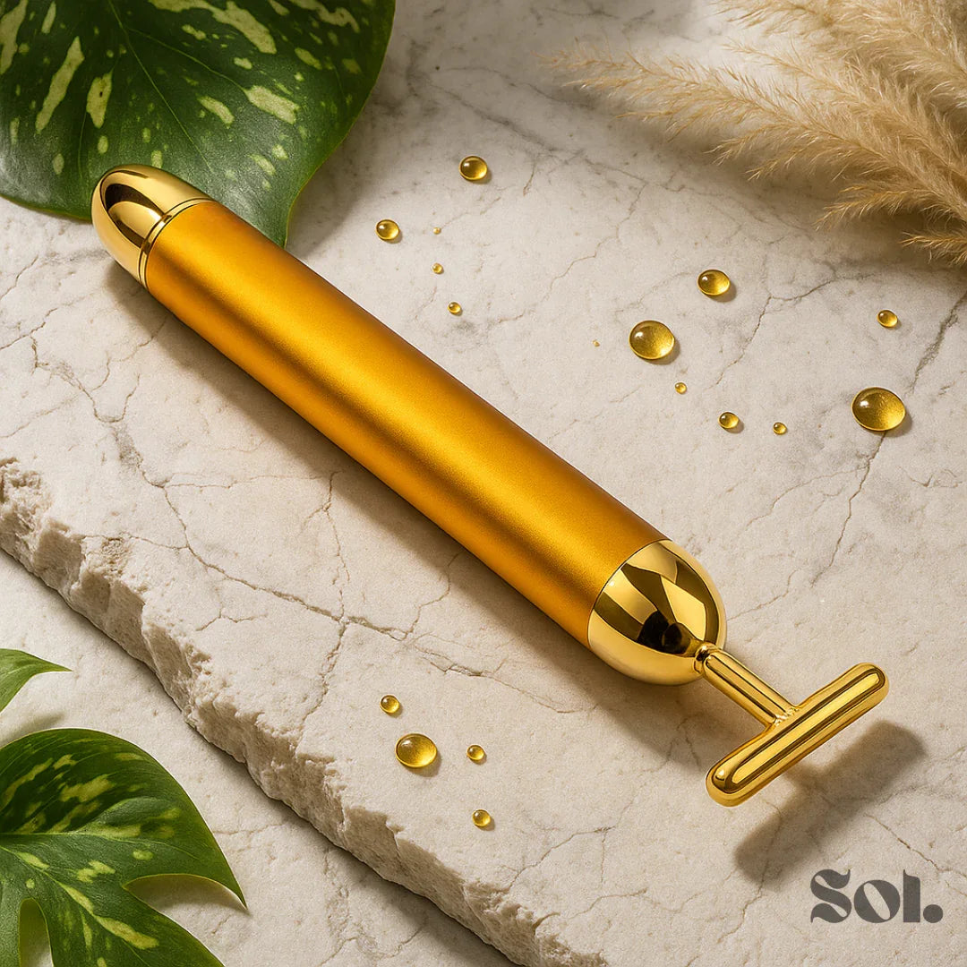 Gold facial massager tool on stone surface with water droplets and green leaves