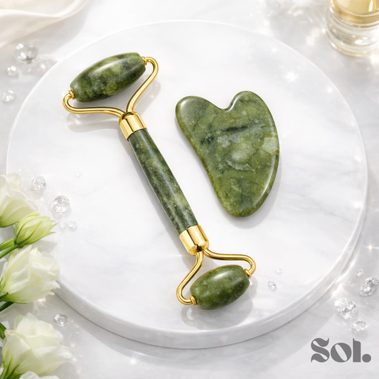 Green jade face roller and gua sha tool on white marble plate with flowers and crystals